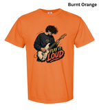 An orange short-sleeve crewneck t-shirt featuring a graphic of a man playing an electric guitar with the text 'PLAY IT LOUD' in a stylized font. The text 'Burnt Orange' is written in black at the top right corner.