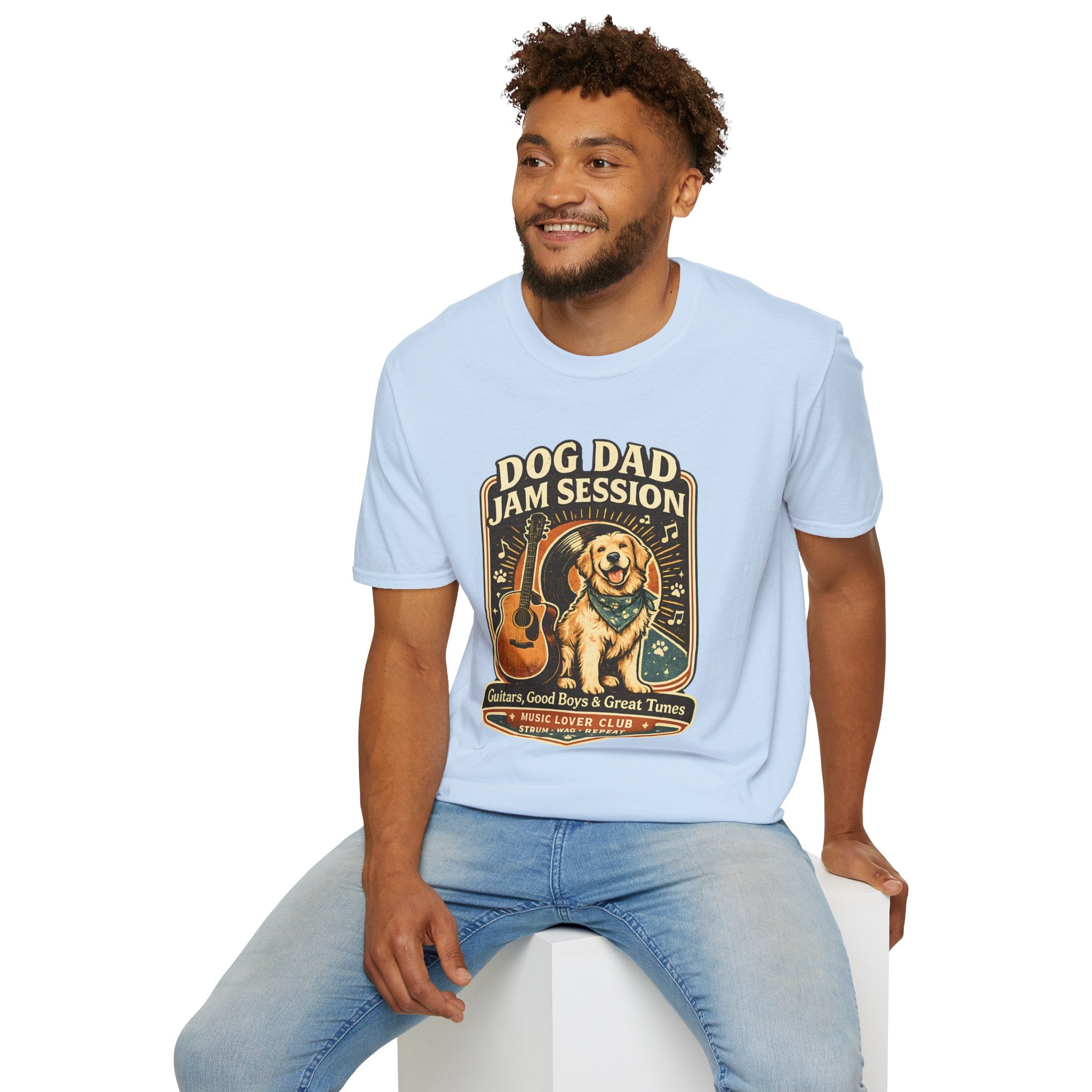 A man wearing a light blue crewneck t-shirt with a 'Dog Dad Jam Session' graphic print, paired with light-wash blue jeans, sitting on a white block against a white background.