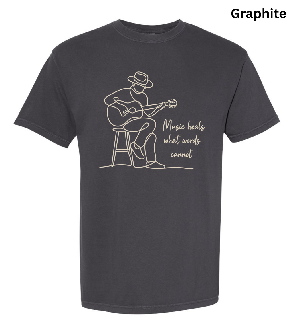 A dark charcoal gray t-shirt with a white line art graphic of a man playing an acoustic guitar on a stool, next to the quote 'Music heals what words cannot.' The word 'Graphite' is printed in black in the top right corner, indicating the fabric color.