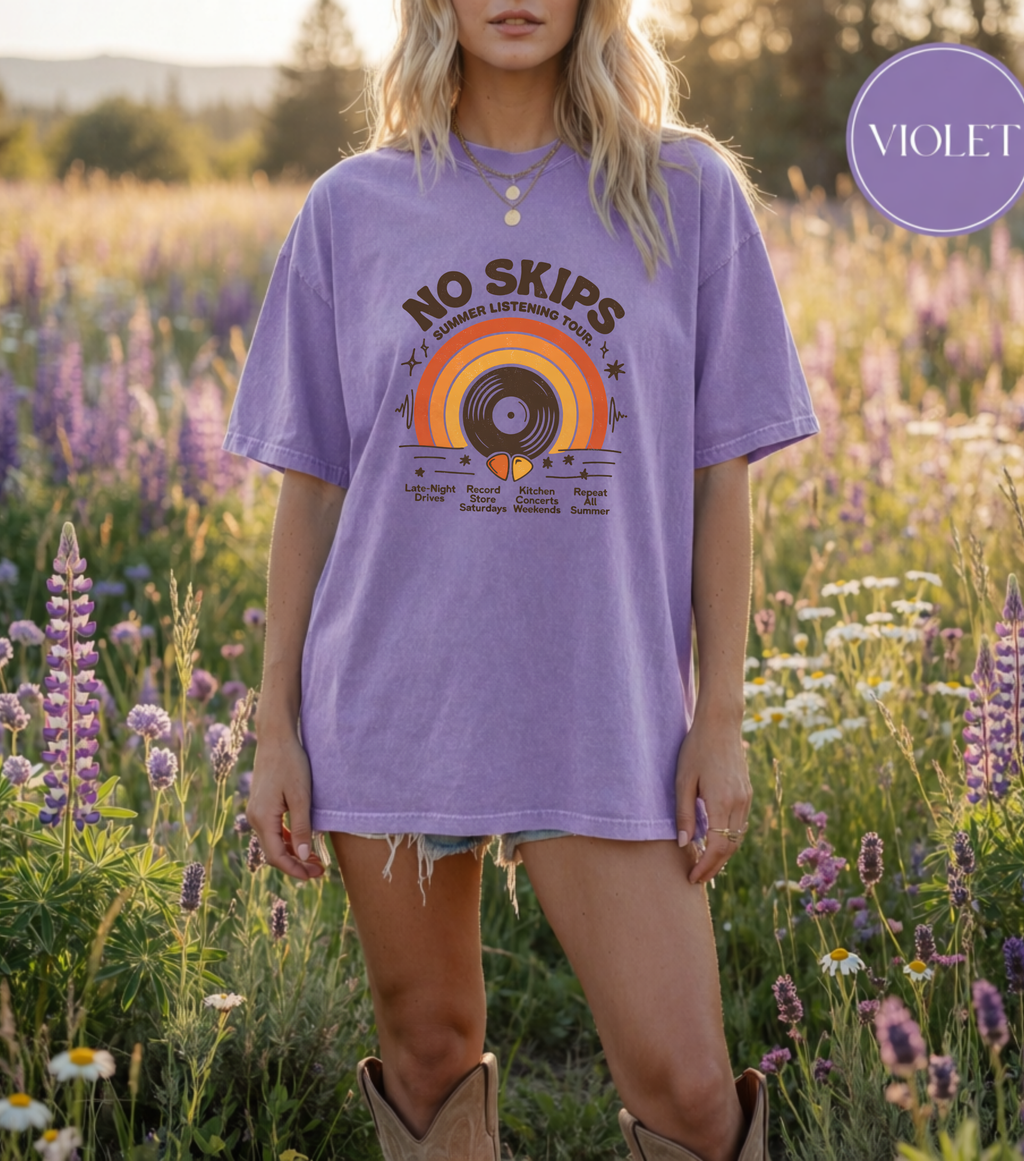 A woman wearing an oversized violet graphic T-shirt stands in a sunlit field of purple flowers. The shirt has a retro vinyl record and sunset print with the text 'NO SKIPS' and smaller text below.
