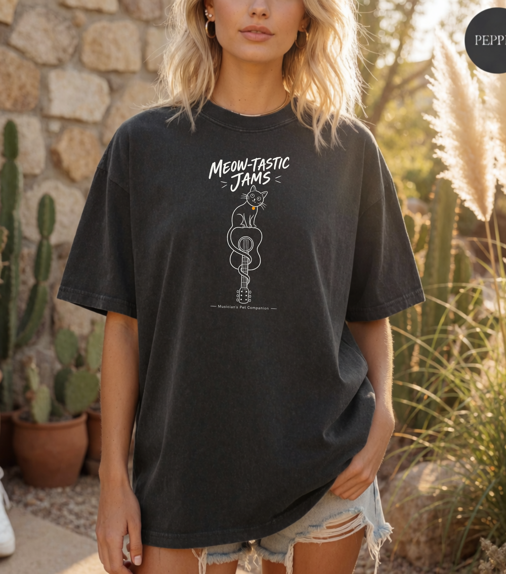 A woman wearing a faded black graphic t-shirt with a white snake coiled around a guitar and the text 'MEOW-TASTIC JAMS'. The background is a warm, sunny outdoor desert setting with cacti and a stone wall.