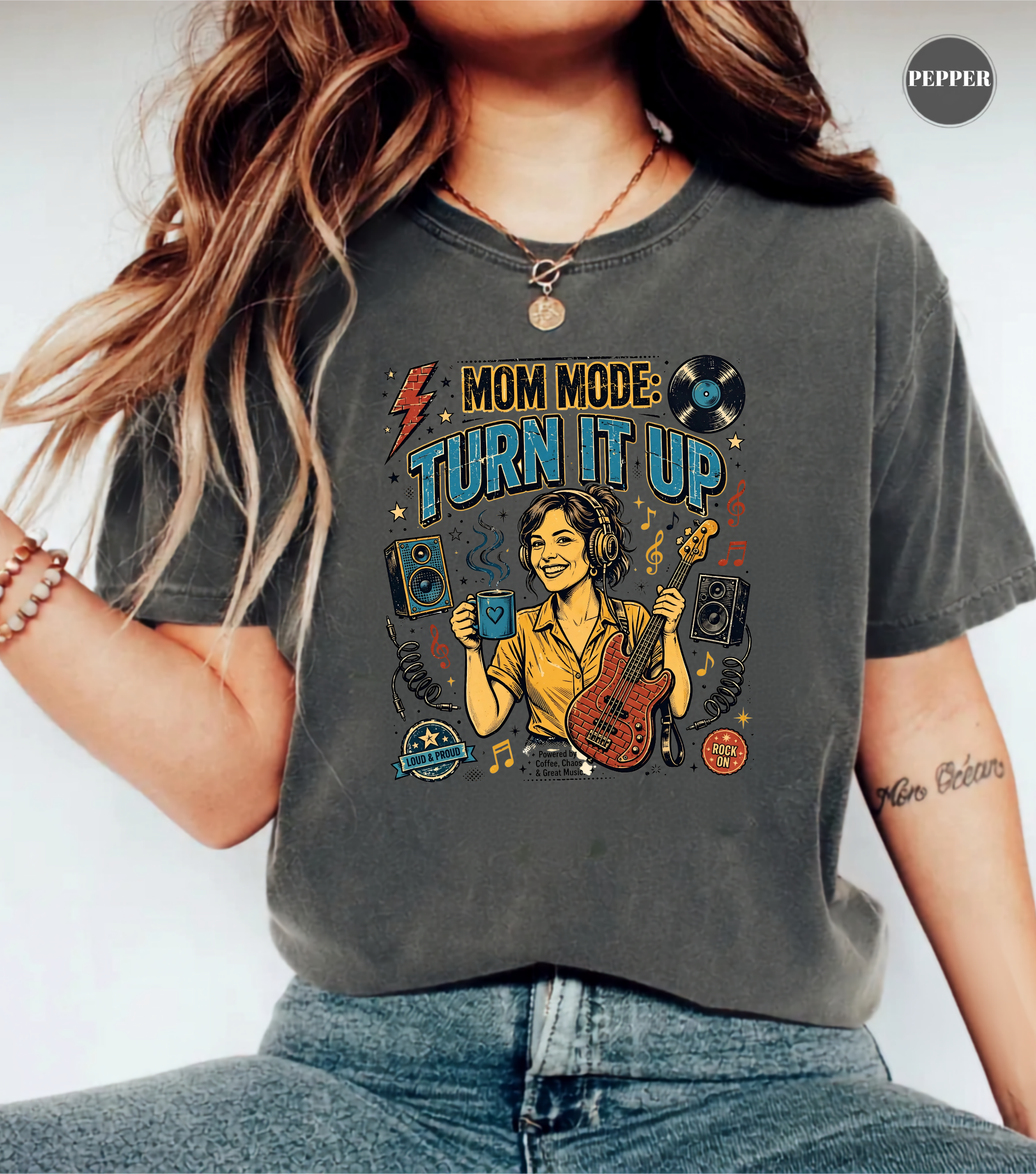 A woman wearing a dark gray graphic t-shirt with a colorful 'MOM MODE: TURN IT UP' design featuring a woman playing guitar and speakers, paired with blue jeans.