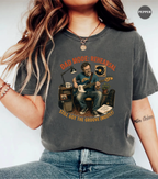 A woman wearing a charcoal gray graphic t-shirt with a vintage-style illustration of a man playing an electric guitar and singing into a microphone. The text on the shirt reads 'DAD MODE: REHEARSAL' and 'STILL GOT THE GROOVE (MOSTLY)'.