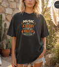 A woman wearing a dark charcoal gray graphic t-shirt with the text 'MUSIC ISN'T A HOBBY IT'S THE WAY I BREATHE' in orange and cream lettering, standing outdoors in front of a stone wall and cacti.