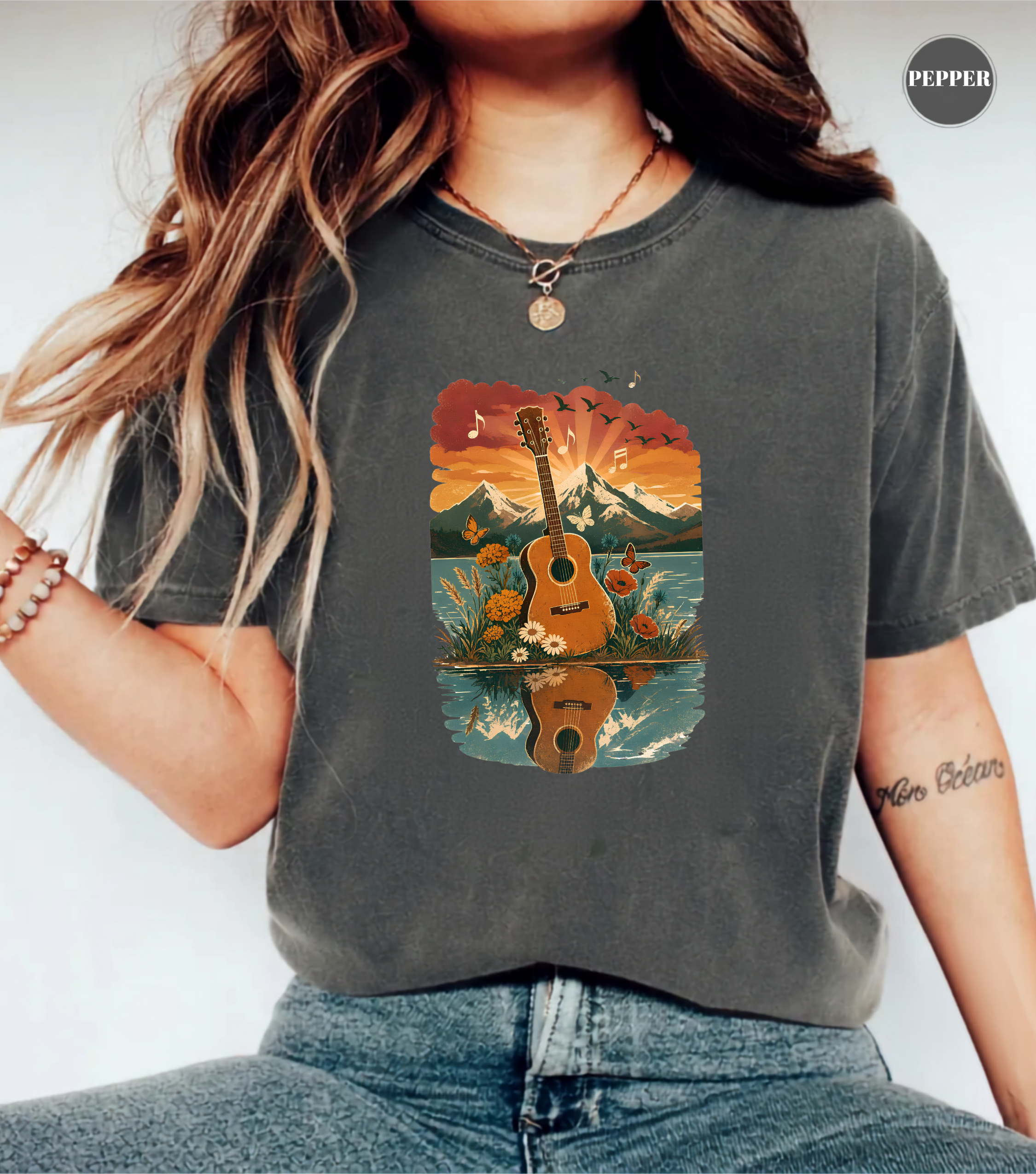 A woman wearing a dark gray graphic t-shirt with a colorful print of an acoustic guitar in front of a mountain lake landscape. The t-shirt has a faded wash texture, and the model is also wearing blue jeans and a necklace.