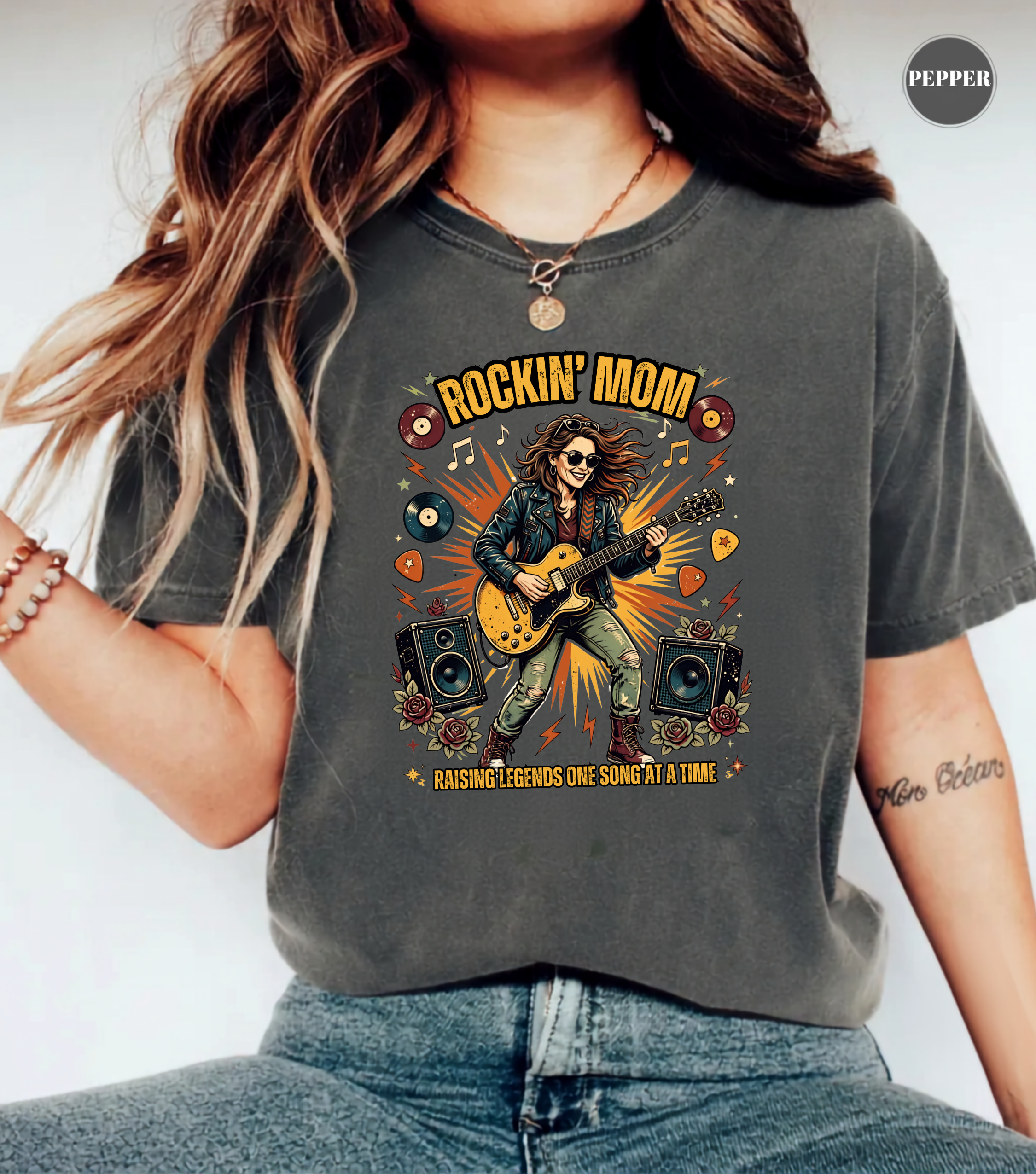 A woman wearing a charcoal gray graphic t-shirt with a 'Rockin' Mom' guitar player design, paired with blue jeans. The t-shirt features a colorful retro graphic of a woman playing an electric guitar with speakers and vinyl records.