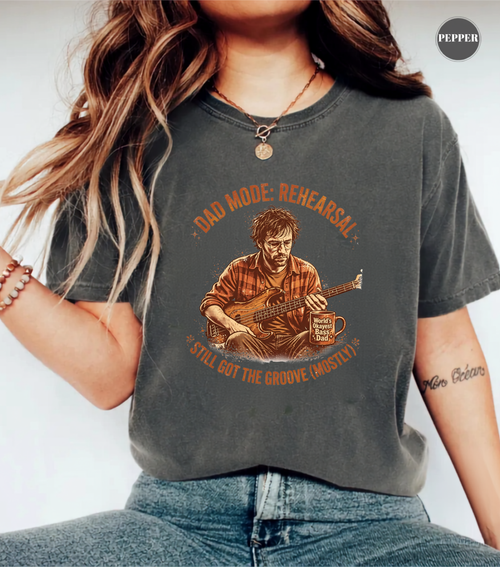 A woman wearing a dark gray graphic t-shirt with a retro-style illustration of a man playing guitar and the text 'DAD MODE: REHEARSAL' and 'STILL GOT THE GROOVE MOSTLY'.
