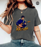 A woman wearing a dark gray graphic t-shirt with a cartoon illustration of a man playing an electric guitar and the text 'PLAY IT LOUD' in yellow. The t-shirt has a washed, vintage texture.