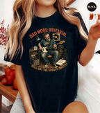 A woman wearing a black graphic t-shirt with a vintage-style illustration of a man playing an electric guitar and singing into a microphone. The text on the shirt reads 'DAD MODE: REHEARSAL' and 'STILL GOT THE GROOVE (MOSTLY!)'.