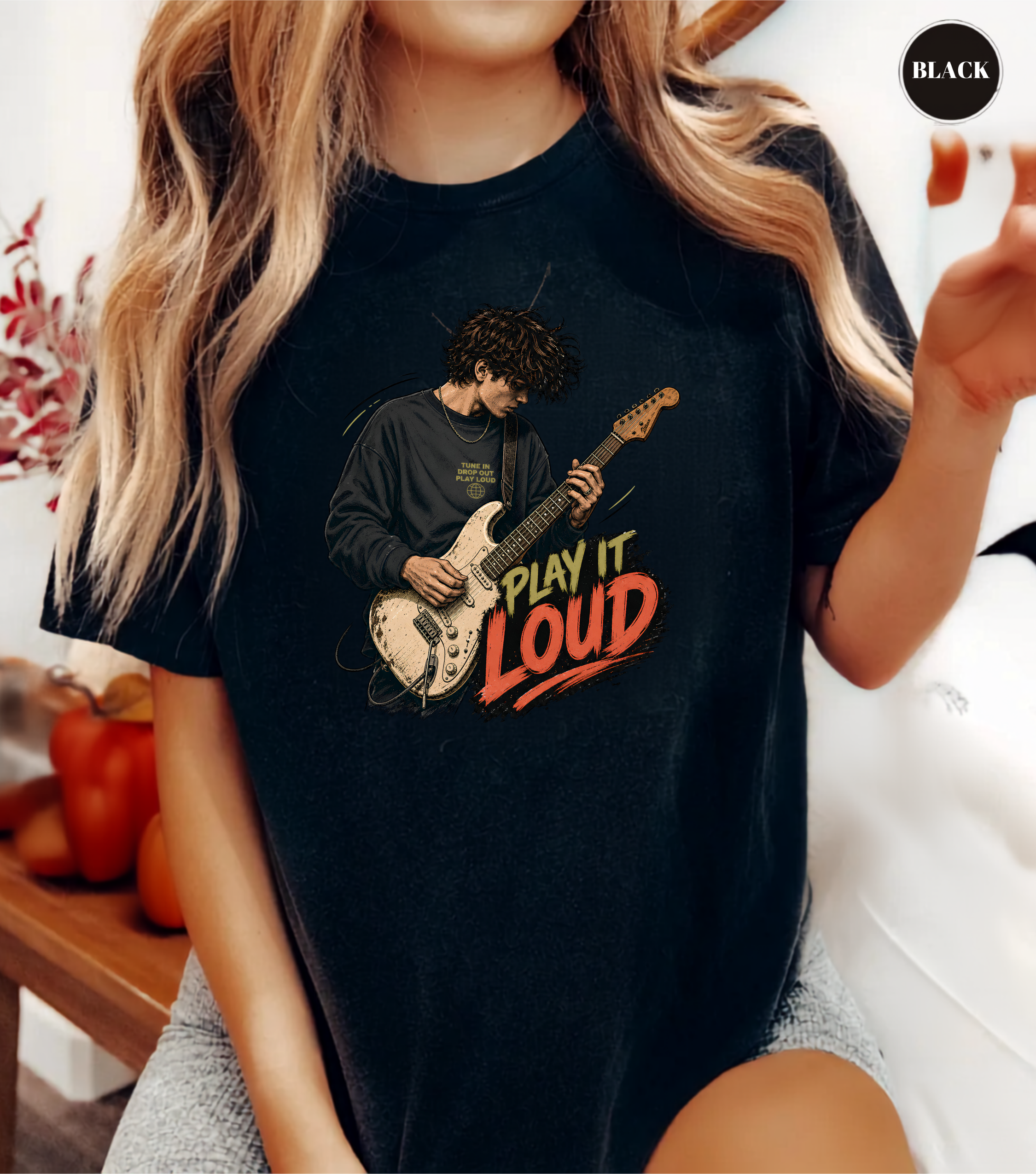 A woman wearing a black graphic t-shirt with a printed illustration of a guitarist and the text 'PLAY IT LOUD' in yellow and red. The image is a lifestyle shot with a 'BLACK' label in the top right corner.