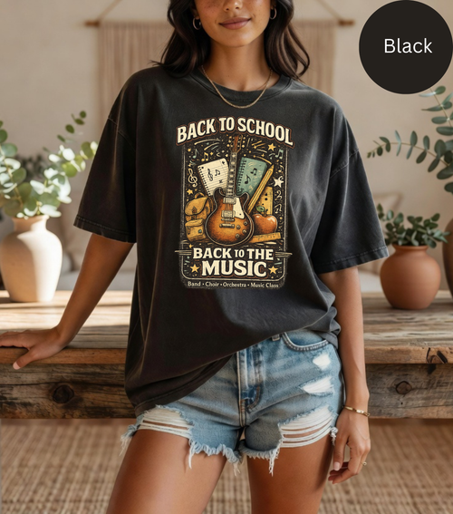 A woman wearing a black graphic t-shirt with a 'Back to School Back to the Music' design featuring guitars and playing cards, paired with distressed denim shorts in a warm indoor setting.