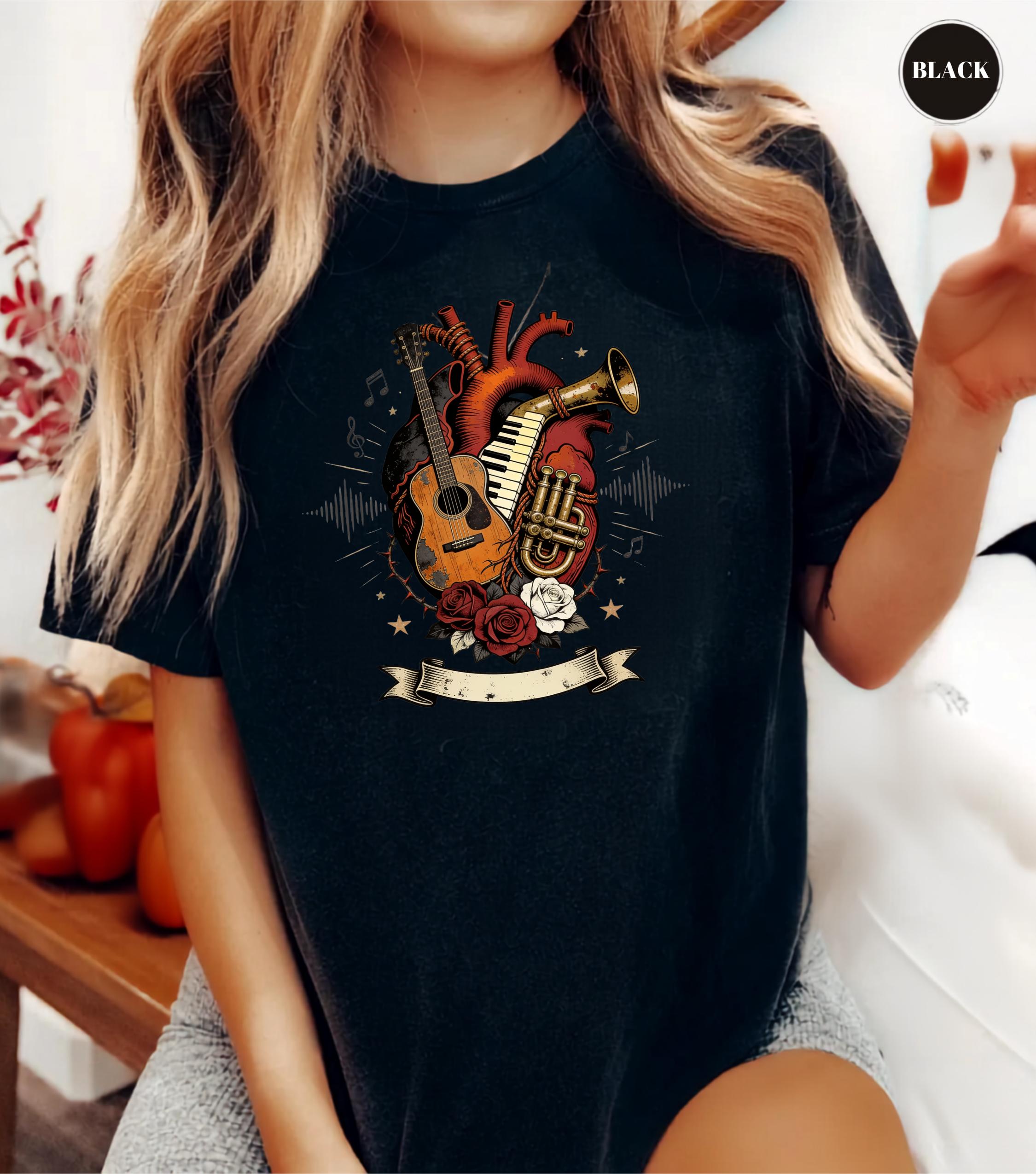 A woman wearing a black graphic t-shirt featuring an anatomical heart design with musical instruments like a guitar, trumpet, and piano keys, set against a lifestyle background with a 'BLACK' label in the top right corner.