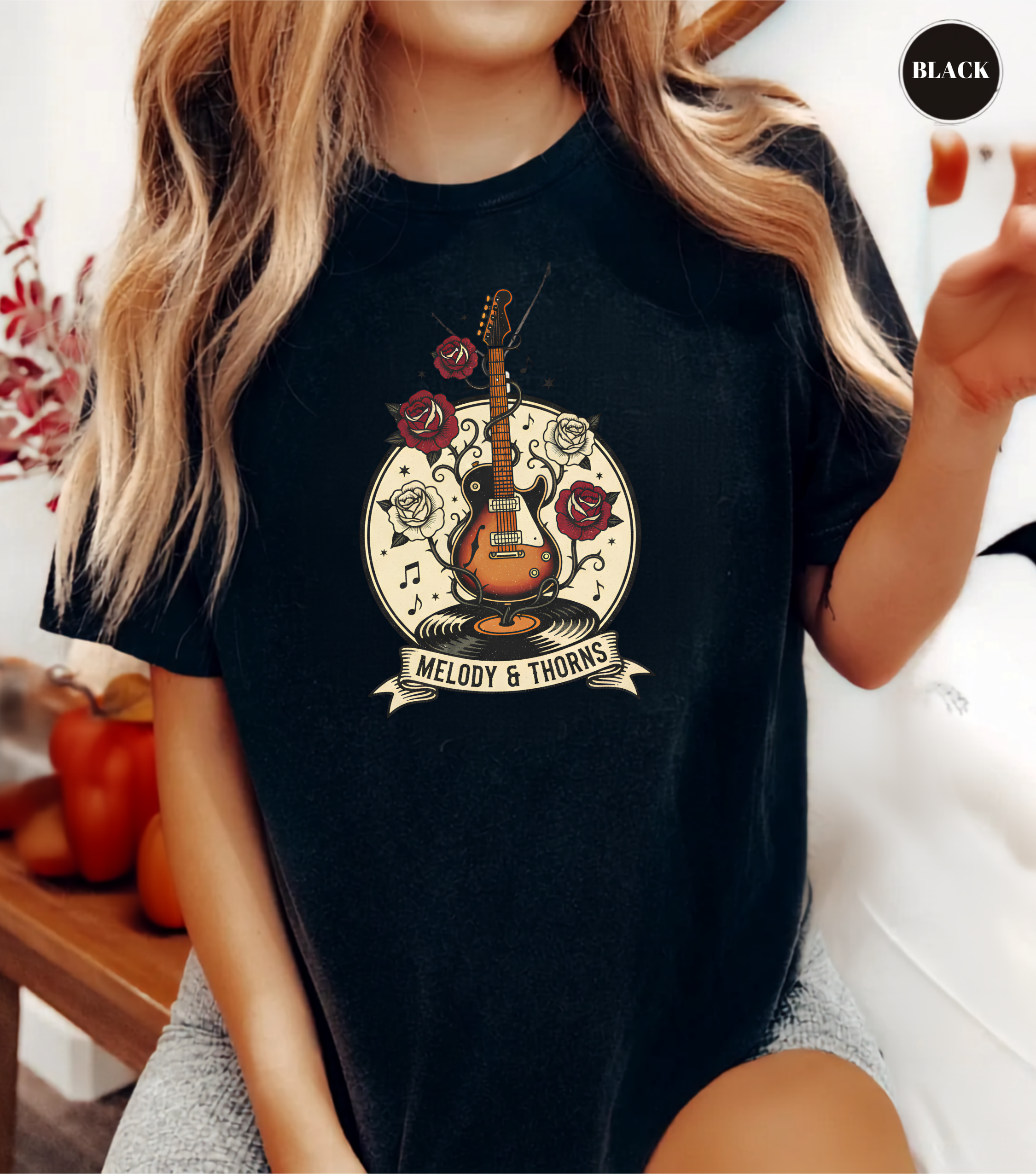 A woman wearing a black graphic t-shirt with a vintage-style guitar, roses, and musical notes design that reads 'Melody & Thorns'.