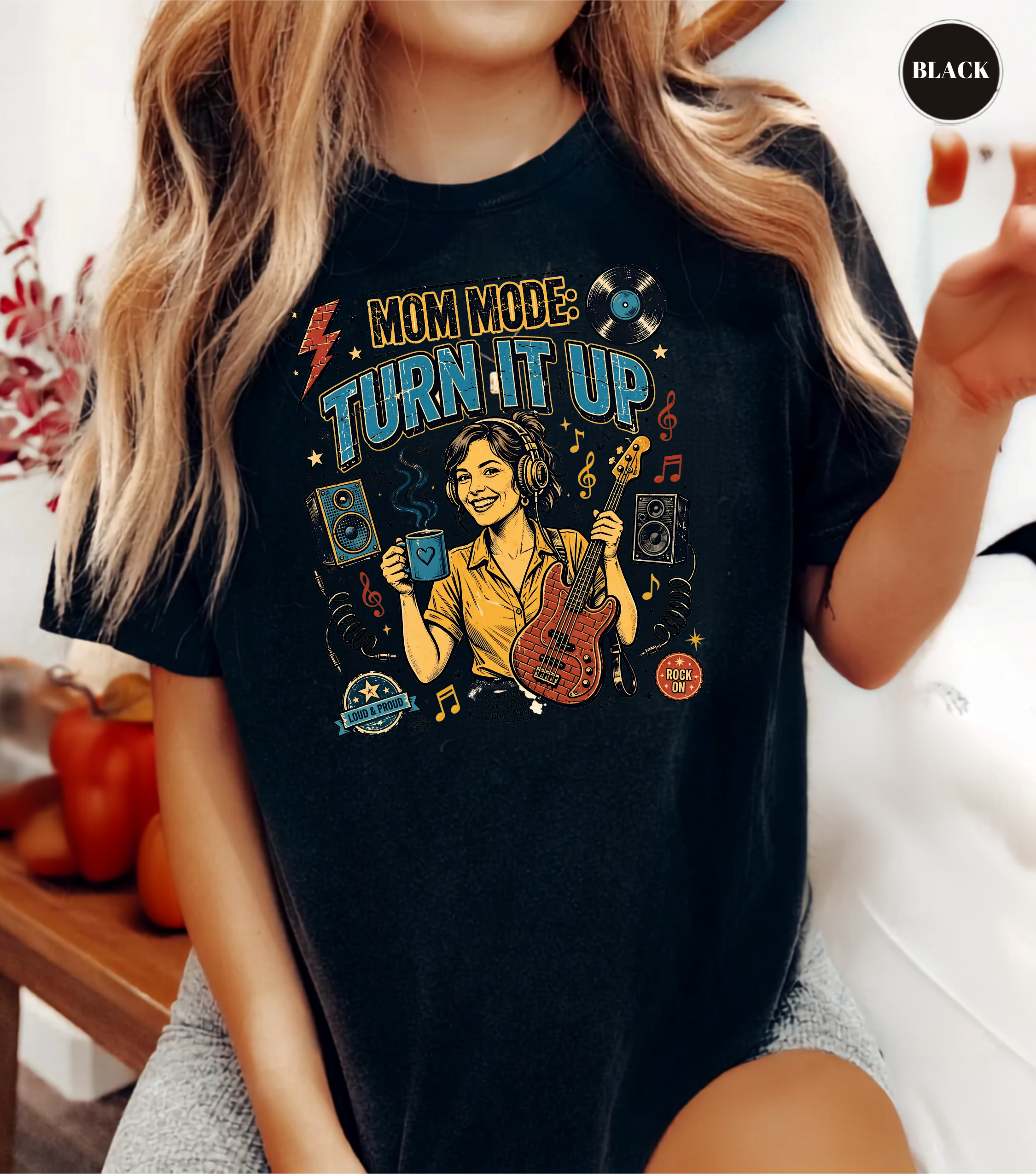 A woman wearing a black graphic t-shirt with a colorful 'Mom Mode: Turn It Up' design featuring a cartoon woman playing guitar and holding a drink. The image has a 'BLACK' label in the top right corner.