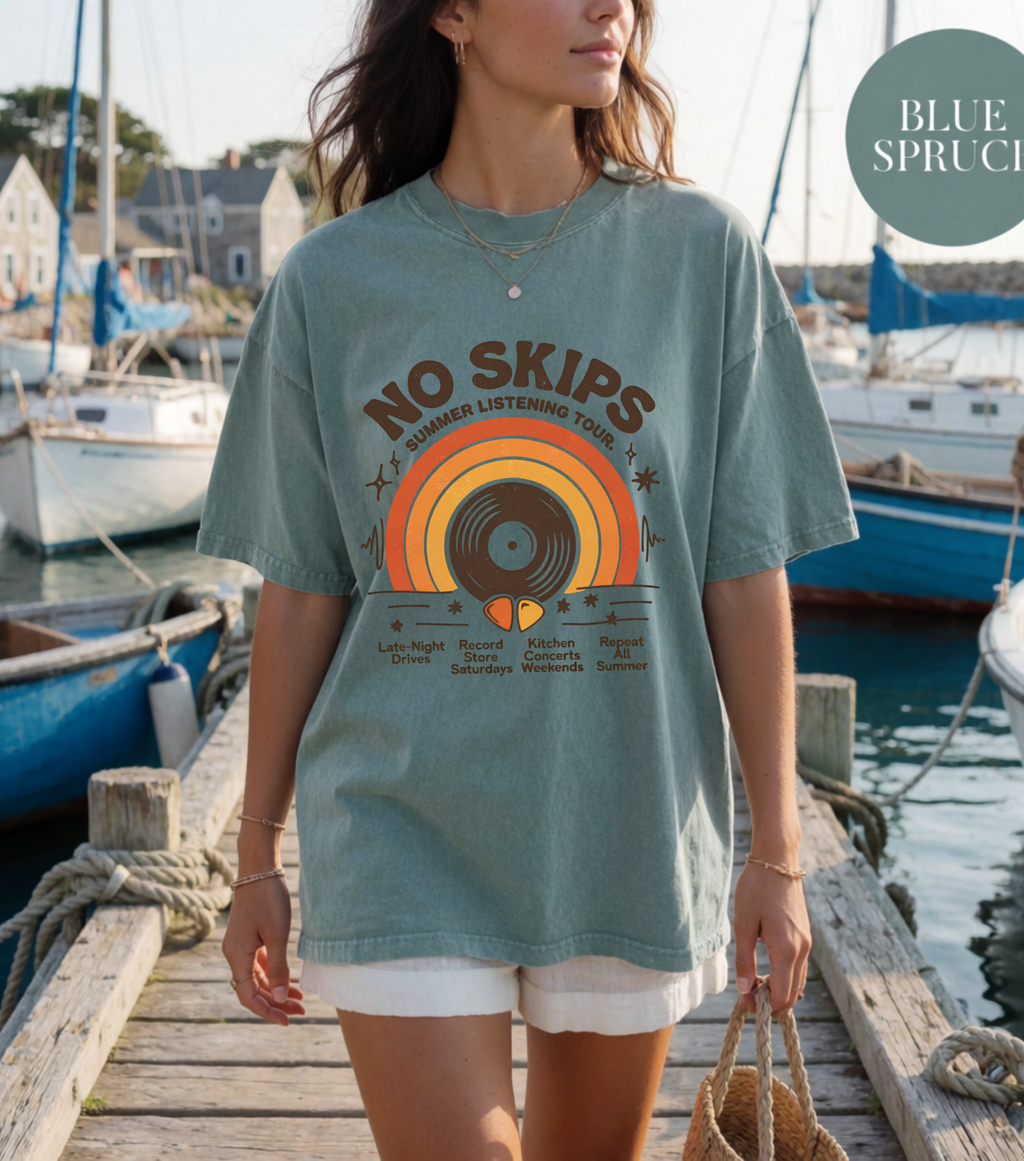 A woman wearing an oversized dusty blue graphic t-shirt on a wooden dock with boats in the background. The t-shirt features a retro vinyl record design with the text 'NO SKIPS SUMMER LISTENING TOUR, N.