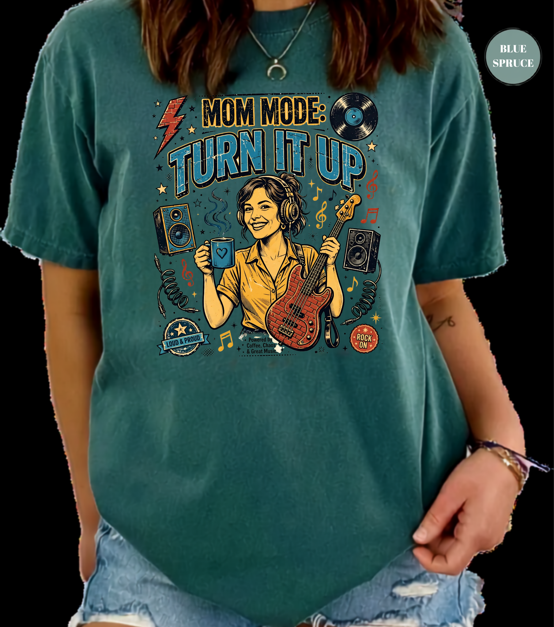 A woman wearing a teal graphic t-shirt with a 'Mom Mode: Turn It Up' design featuring a cartoon bassist and music icons, paired with denim shorts against a black background.