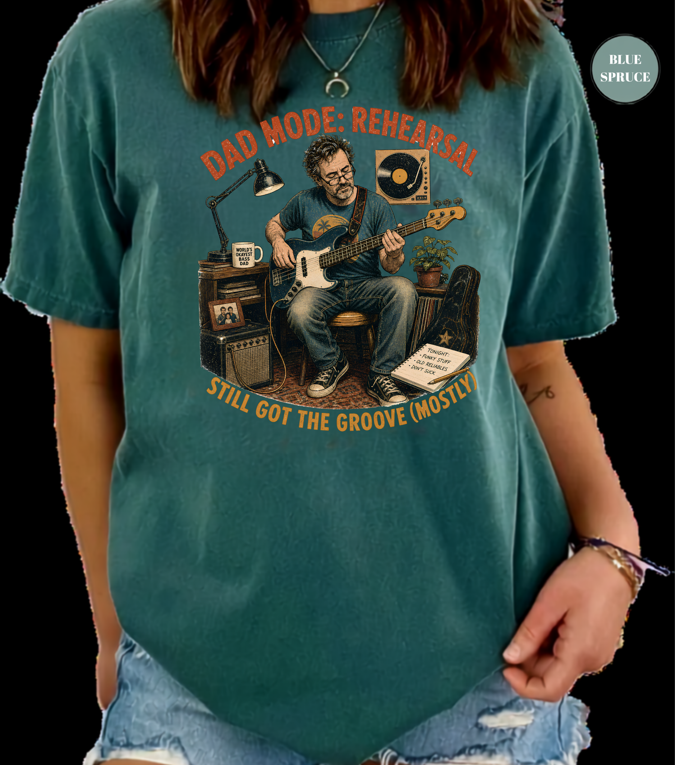 A person wearing a teal graphic T-shirt with a vintage-style illustration of a man playing guitar and the text 'DAD MODE: REHEARSAL' and 'STILL GOT THE GROOVE (MOSTLY)'. The photo is cropped from the neck down against a black background, with a small 'BILL SPRUCE' label in the top right corner.