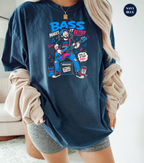 A woman wearing an oversized navy blue graphic t-shirt with a colorful cartoon bass player design and the text 'BASS MAKES IT BETTER'. She has a beige corduroy shirt draped off her shoulders and gray shorts, standing in a brightly lit indoor setting.