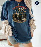 A woman wearing an oversized navy blue graphic t-shirt with a vintage-style illustration of a man playing guitar and the text 'DAD MODE: REHEARSAL' and 'STILL GOT THE GROOVE (MOSTLY)'. She has a beige ribbed sweater draped off her shoulders and gray shorts, set against a bright, minimalist indoor background.
