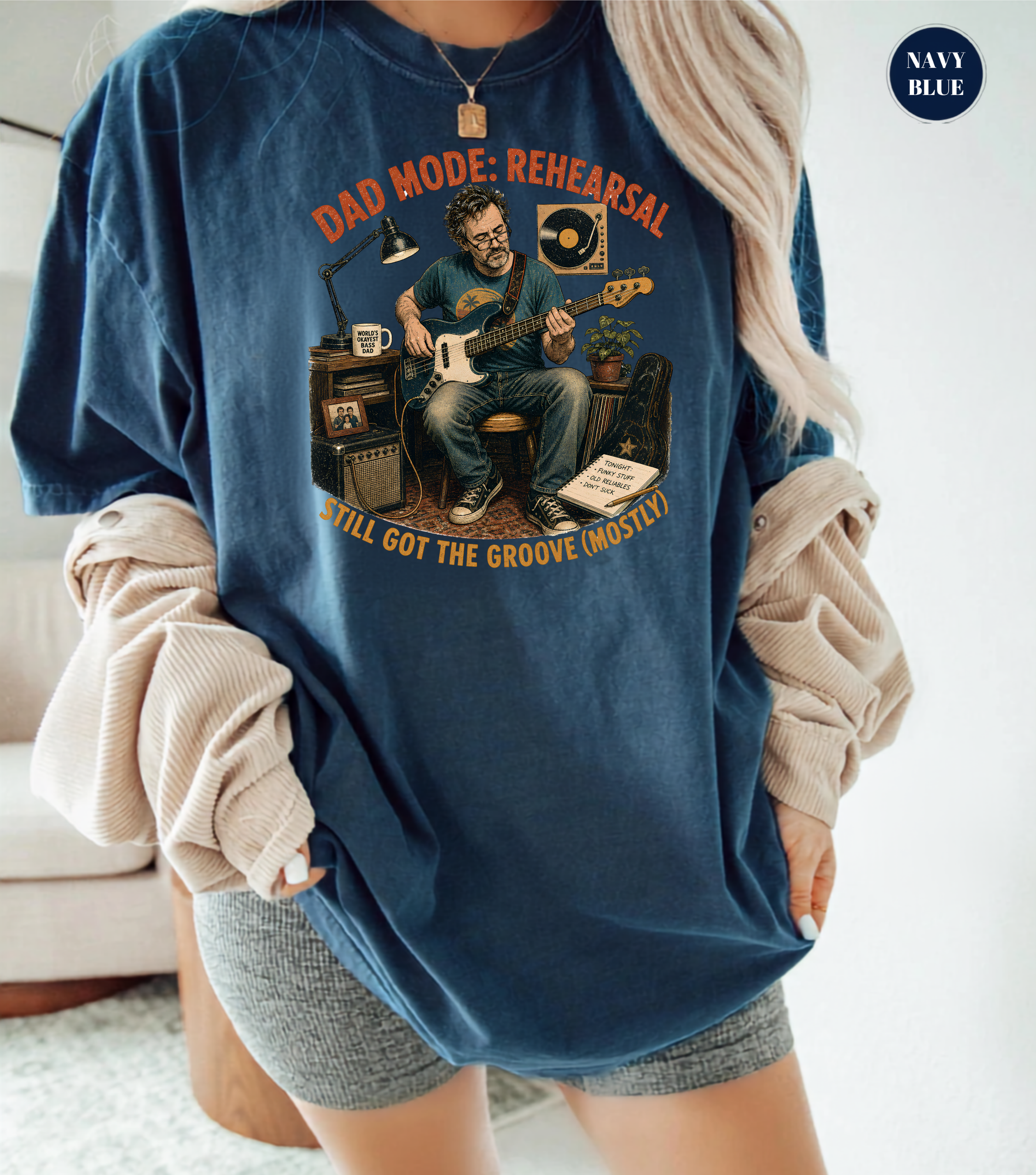 A woman wearing an oversized navy blue graphic t-shirt with a vintage-style illustration of a man playing guitar and the text 'DAD MODE: REHEARSAL' and 'STILL GOT THE GROOVE (MOSTLY)'. She has a beige ribbed sweater draped off her shoulders and gray shorts, set against a bright, minimalist indoor background.