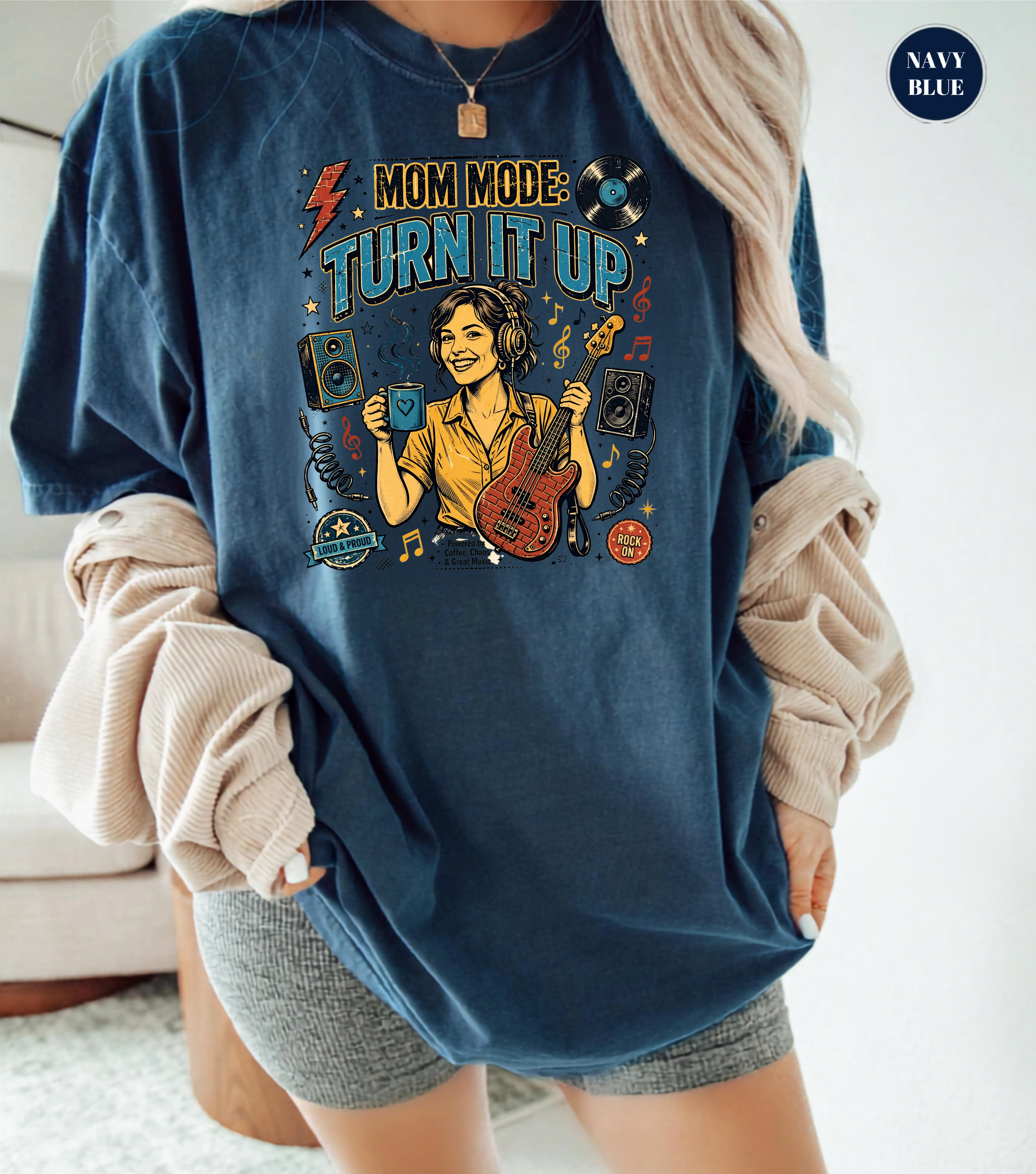 A woman wearing an oversized navy blue graphic t-shirt with a 'Mom Mode: Turn It Up' design, styled with beige rolled-up long sleeves and gray shorts.