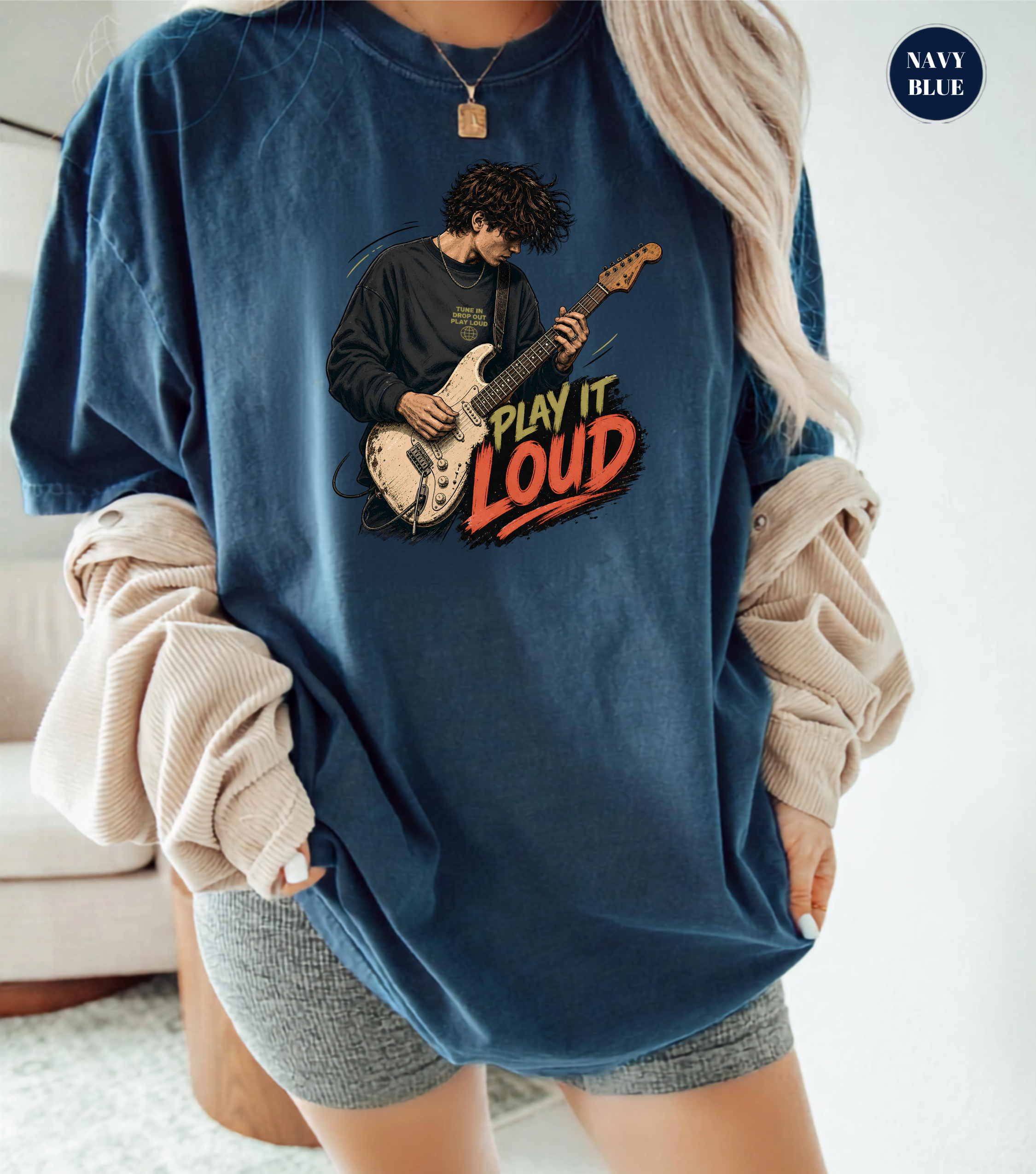 A woman wearing an oversized navy blue graphic t-shirt with a guitar player illustration and the words 'PLAY IT LOUD' in red and orange. She has beige ribbed long sleeves draped off her shoulders and is wearing gray shorts.
