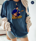 A woman wearing an oversized navy blue graphic t-shirt with a cartoon illustration of a man playing guitar and the text 'PLAY IT LOUD'. She has beige ribbed knit sleeves draped off her shoulders and is wearing gray shorts.