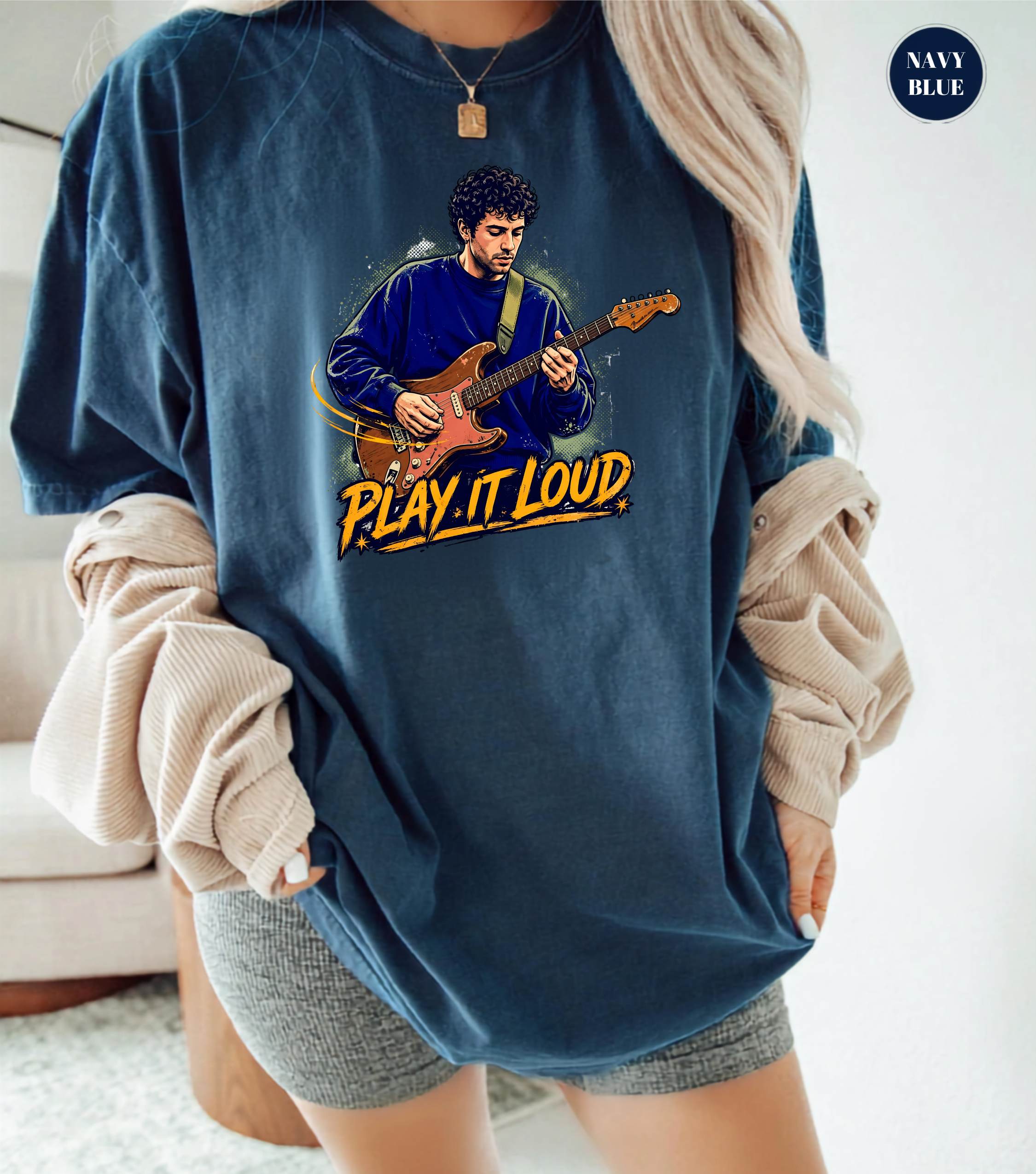 A woman wearing an oversized navy blue graphic t-shirt with a cartoon illustration of a man playing guitar and the text 'PLAY IT LOUD'. She has beige ribbed knit sleeves draped off her shoulders and is wearing gray shorts.