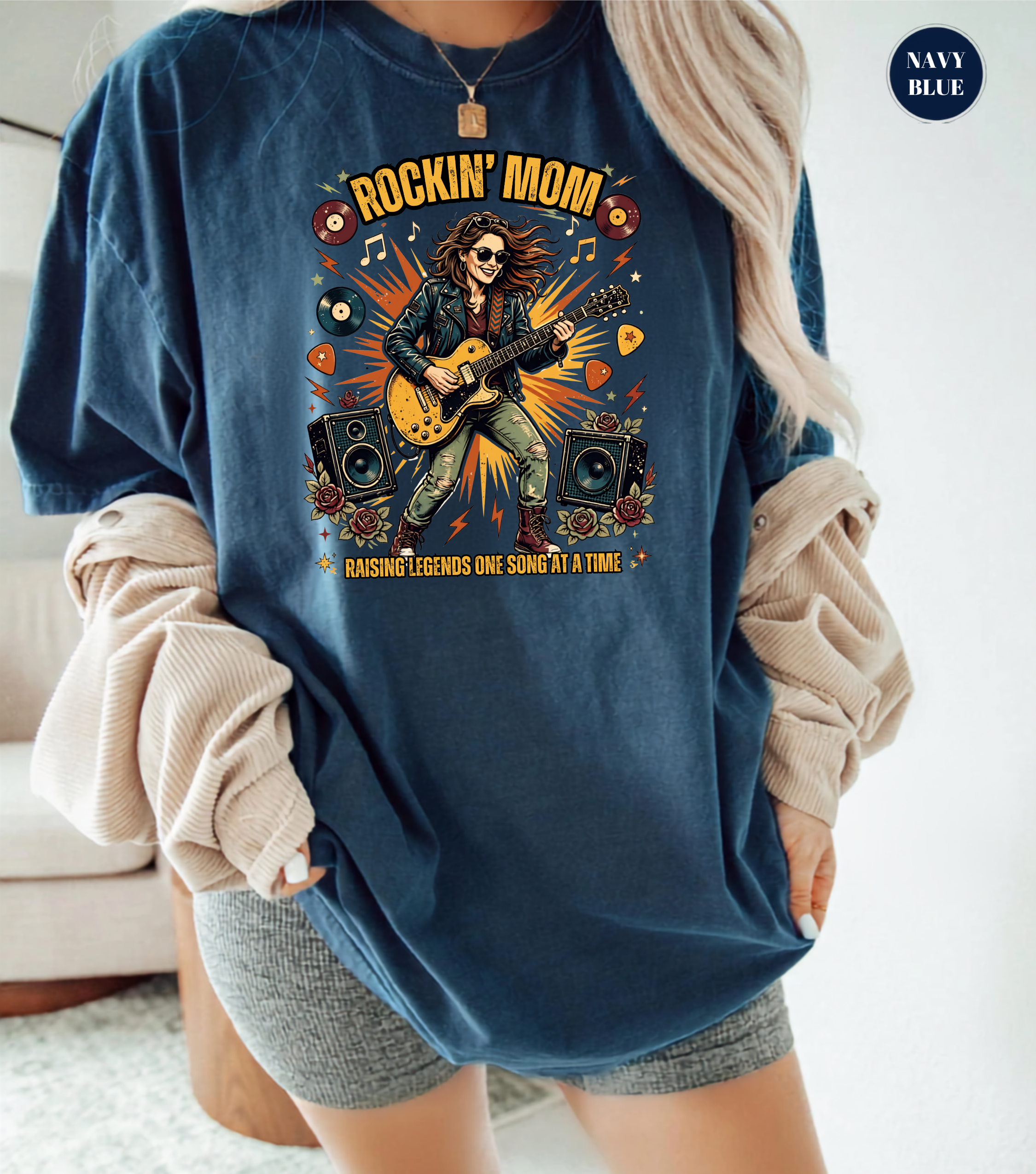 A woman wearing an oversized navy blue graphic T-shirt with a colorful rockstar guitar design and the words 'ROCKIN' MOM' and 'RAISING LEGENDS ONE SONG AT A TIME.' She has beige ribbed long sleeves draped off her shoulders and gray shorts, standing indoors near a couch.