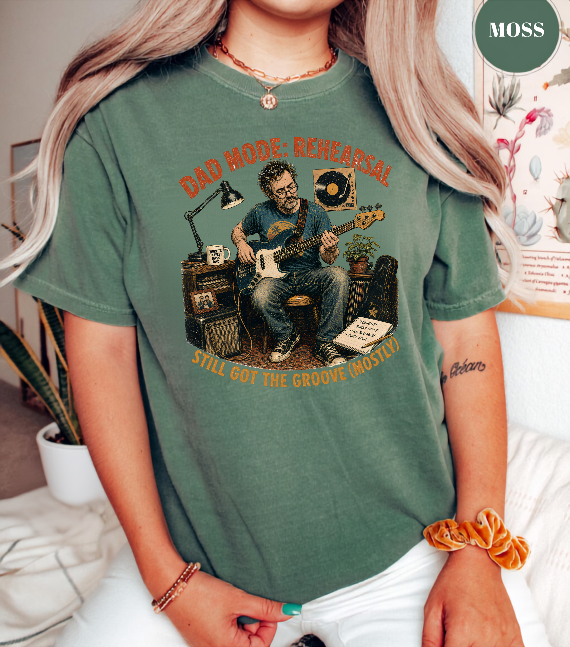A woman wearing a sage green graphic t-shirt with a vintage-style illustration of a man playing an electric guitar in a rehearsal room. The text on the shirt reads 'DAD MODE, REHEARSAL' and 'STILL GOT THE GROOVE GHOSTS'.