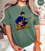 A woman wearing a heathered sage green graphic t-shirt with a printed illustration of a man playing an electric guitar and the text 'PLAY IT LOUD'. The t-shirt has a 'MOSS' brand label in the top right corner.