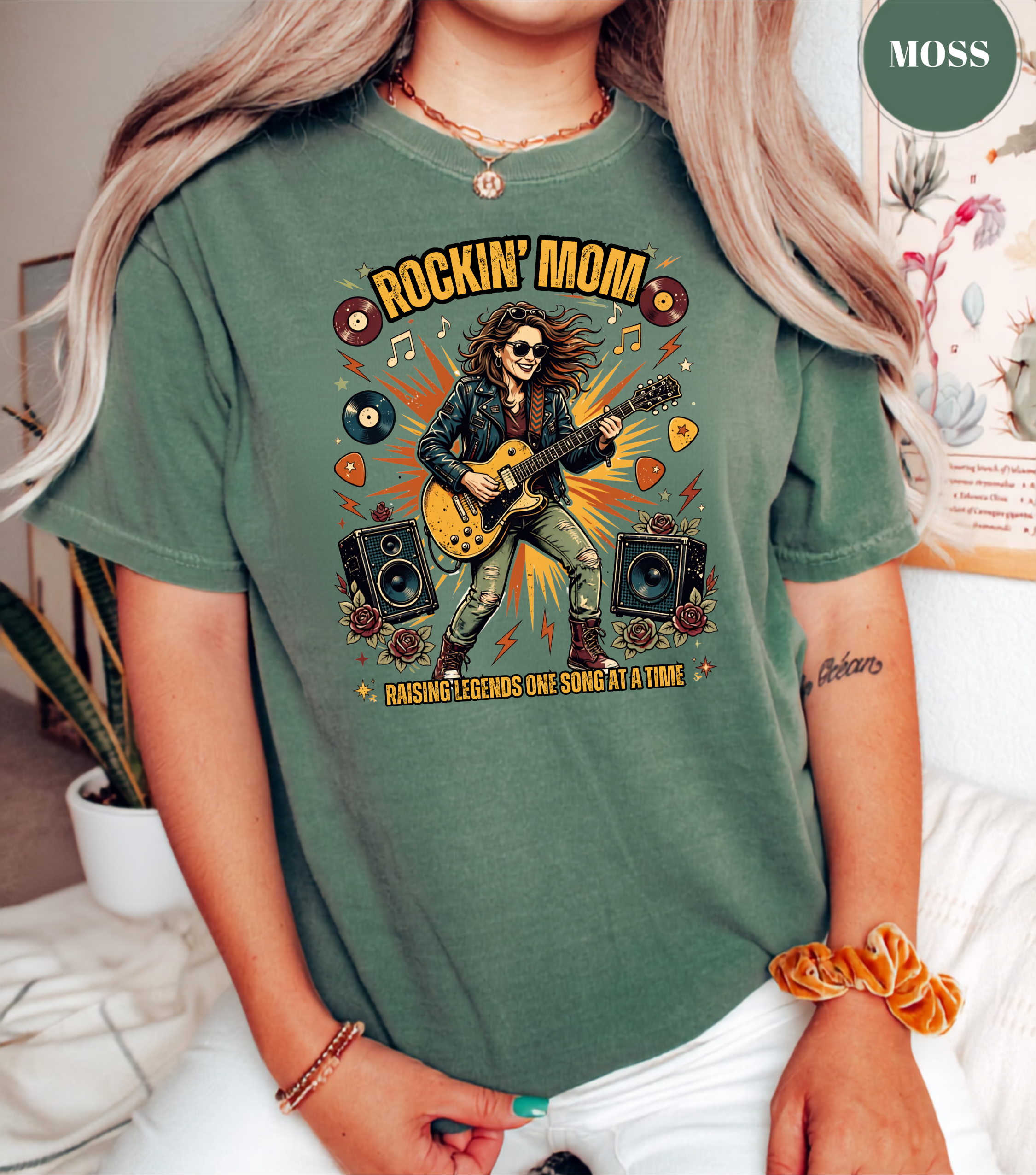 A woman wearing a sage green graphic t-shirt with a 'Rockin' Mom' guitar player design, in a cozy indoor setting.