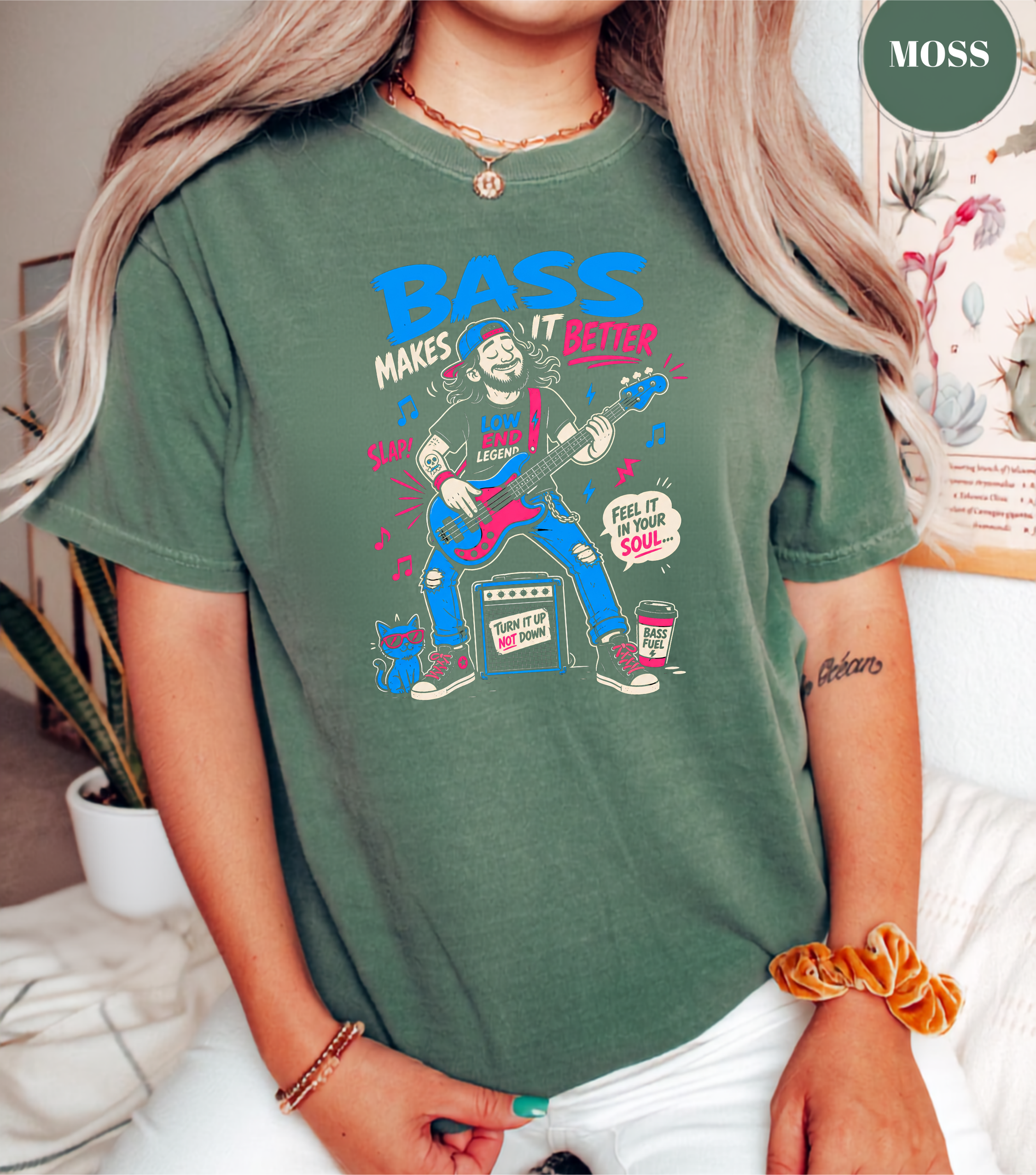 A woman wearing a green graphic t-shirt with a colorful illustration of a girl playing a guitar and the text 'BASS MAKES IT BETTER'. The brand name 'MOSS' is visible in a green circle in the top right corner.