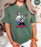 A woman wearing a sage green graphic t-shirt with a colorful illustration of a guitarist and the text 'SLAP HAPPY'. The background shows a cozy indoor setting with a plant and framed art.