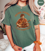 A woman wearing a sage green graphic t-shirt featuring a vintage-style illustration of a man playing a guitar with the text 'BAD MORE, NEW KISHL' and 'GUITAR IS THE ONLY THING I KNOW'.