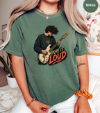 A woman wearing a faded green graphic t-shirt with a printed illustration of a guitarist and the text 'PLAY IT LOUD'. The image has a 'MOSS' brand label in the top right corner.