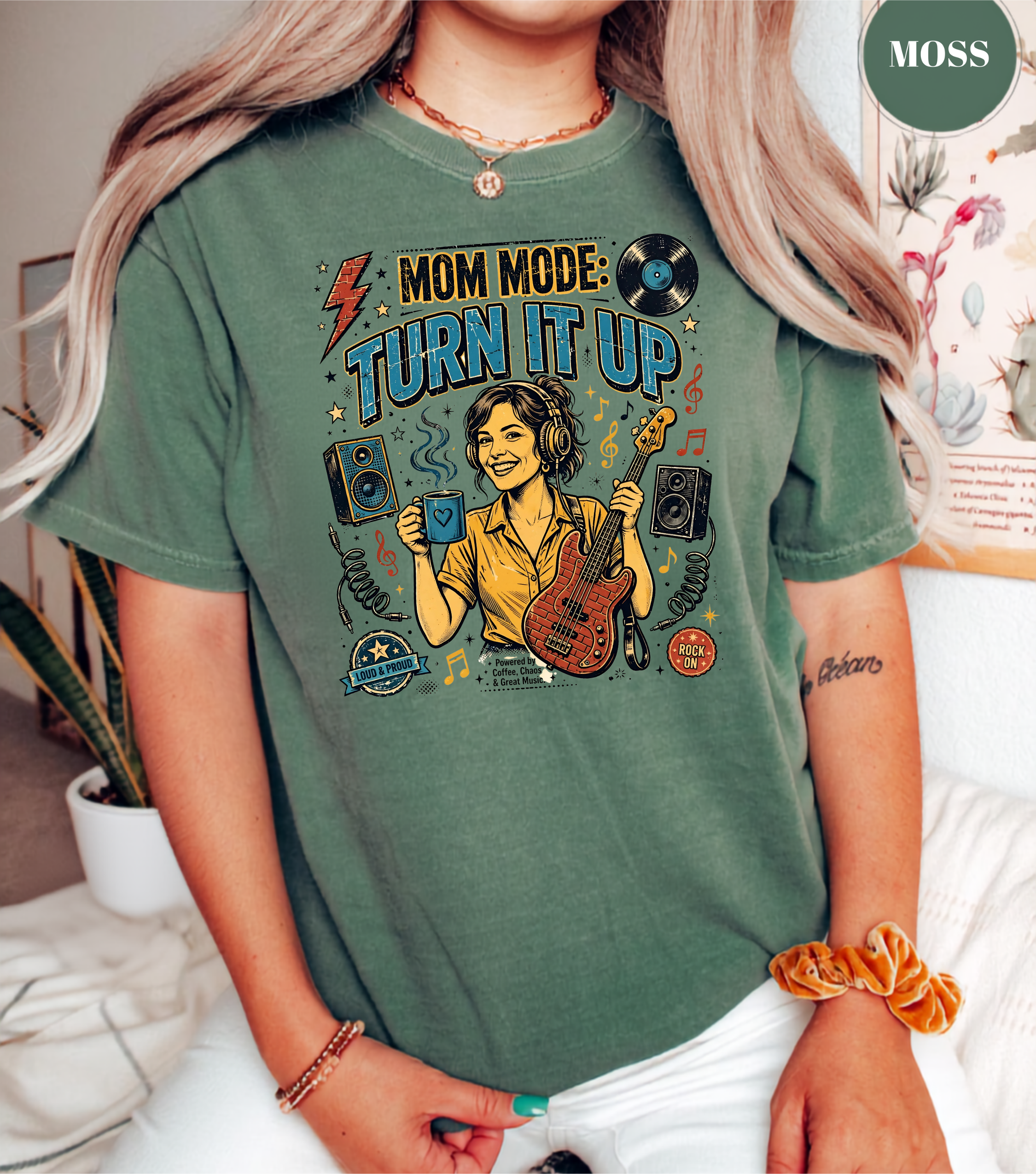 A woman wearing a muted sage green graphic t-shirt with a colorful 'Mom Mode: Turn It Up' design featuring a guitar, speakers, and vinyl records. The brand name 'MOSS' is visible in a circular badge in the top right corner.