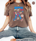A woman wearing a brown graphic t-shirt with a colorful cartoon illustration and the text 'PASS MAKES IT BETTER'. The t-shirt is a washed, faded brown color.