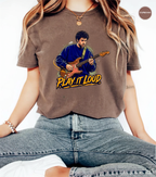 A woman wearing an espresso brown graphic t-shirt with a cartoon illustration of a man playing an electric guitar and the text 'PLAY IT LOUD'.