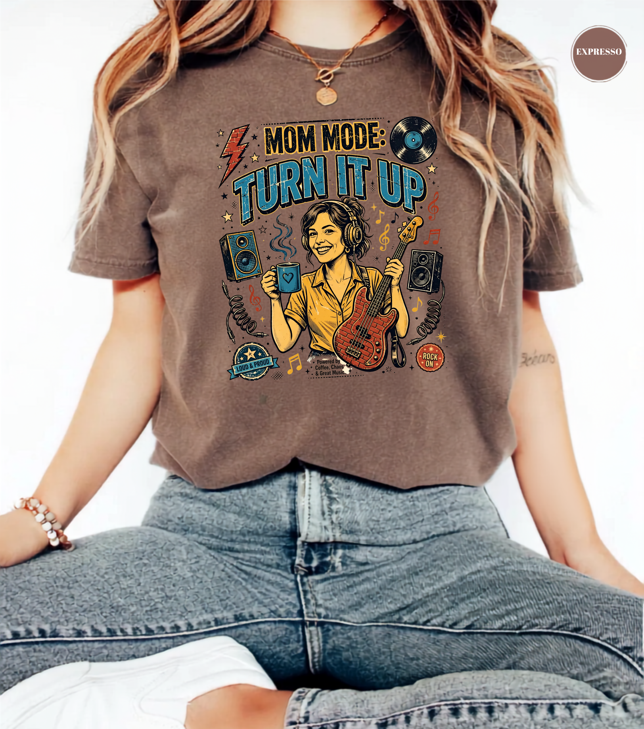 A person wearing a brown graphic T-shirt with the text 'MOM MODE TURN IT UP' and a colorful illustration of a woman DJing, paired with blue jeans against a plain white background.