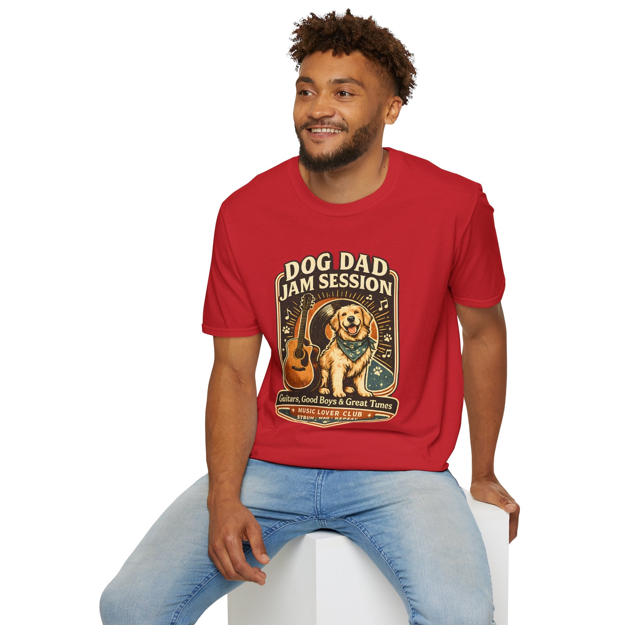 A man wearing a red graphic t-shirt with a 'Dog Dad Jam Session' guitar dog design, paired with light blue jeans, sitting on a white block against a white background.